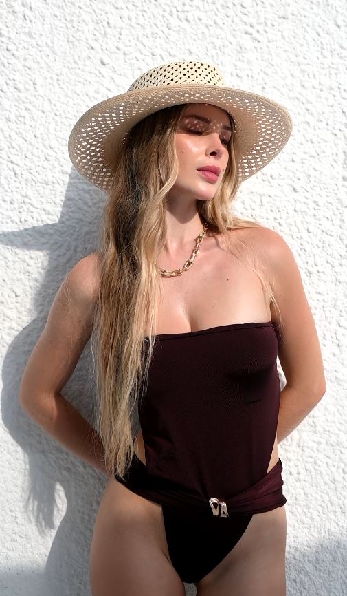 A woman wearing a dark burgundy strapless one-piece swimsuit with a small gold logo detail, a beige woven straw hat, and sunglasses, posing against a white textured wall.